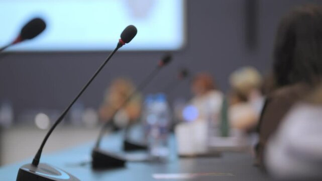 Microphone is on a table with a group of people. The microphone is black and red. The people are sitting at a table with a projector screen behind them