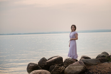 A girl in a long dress stands on the rocks by the lake at sunset