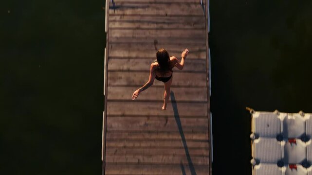 Slow motion shot of a girl jumping into the forest lake from a wooden ponton. Woman running on a wooden pier and jumping into the lake at sunset. Lens flare, top shot, aerial drone shot. Summer vibes.