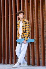 Young man in checkered shirt and light jeans holding skateboard, standing against brick wall and...
