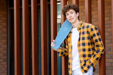 A young man in a yellow checkered shirt stands against a brick wall holding a blue skateboard. His...