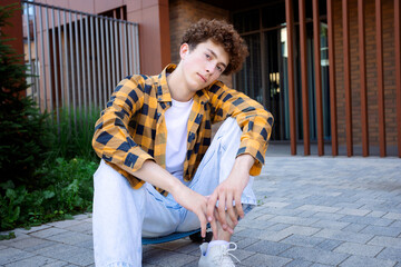 A young man in a checkered shirt sits on a skateboard, looking thoughtfully at the camera. The...
