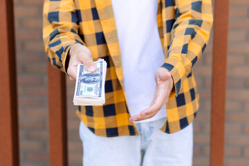 A young man in a checkered shirt holds a wad of dollars while standing near a brick wall. His...