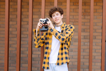 A young man in a yellow and black checked shirt stands against a brick wall holding a vintage...