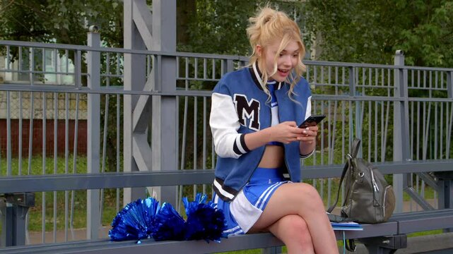 Attractive high school girl in cheerleader uniform and varsity jacket sitting on bleacher, networking and online messaging in social media using cellphone, being surprised and shocked by latest rumors