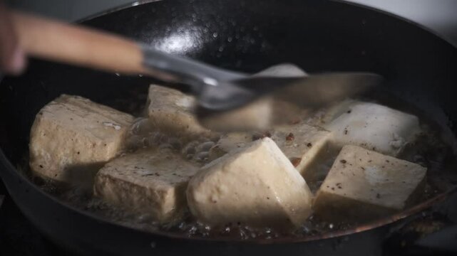 tofu pieces are being flipped in hot oil inside a pan, sizzling as they cook to a golden brown. The motion creates ripples in the bubbling oil, highlighting the frying process. This scene captures. 4K