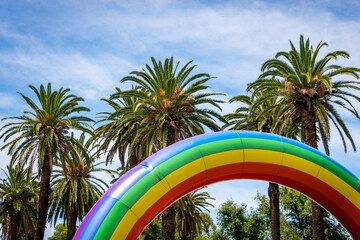 An inflated Pride rainbow against a blue sky and palm trees, horizontal