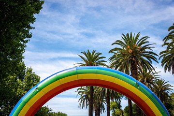 An inflated Pride rainbow against a blue sky and palm trees, symetrical