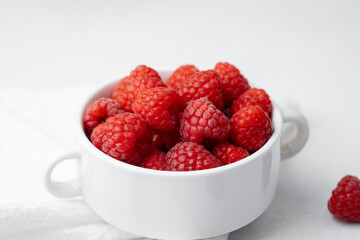 Raspberries in white bowl with scattered berries on light surface for organic food or vegan diet...