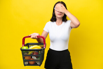 Young woman holding a shopping basket full of food isolated on yellow background covering eyes by...