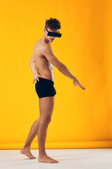 Stylish young man in black shorts posing with a blindfold against a vibrant yellow background,...