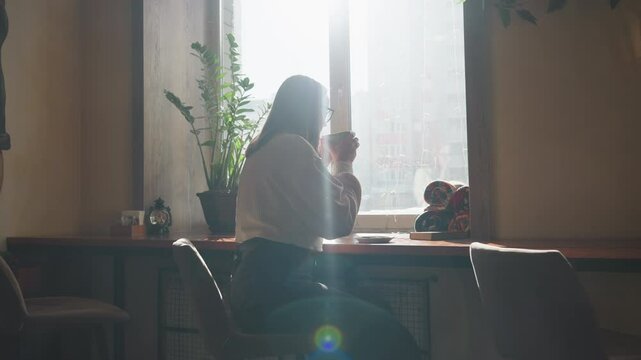 Young girl drinks tea beside window as sunlight streams in, creating dreamy effect, potted plant adds calm touch while distant cityscape shows through glass in quiet, thoughtful atmosphere