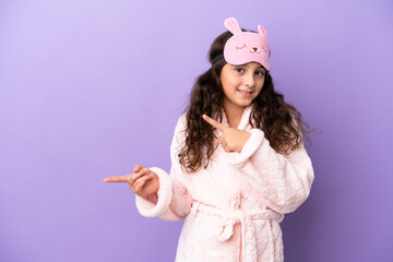 Little caucasian girl in pajamas isolated on purple background surprised and pointing side