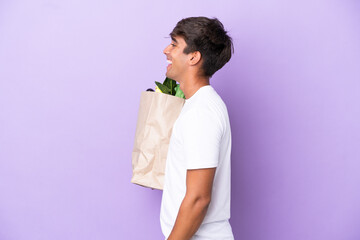 Young man holding a grocery shopping bag isolated on purple background laughing in lateral position