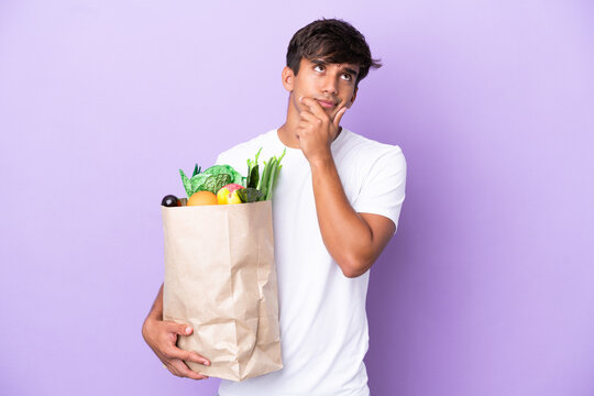 Young man holding a grocery shopping bag isolated on purple background having doubts
