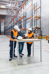 Engineers and manager wearing hardhats discussing logistics over blueprints in warehouse with tall...