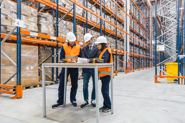 Warehouse workers and manager wearing safety vests and hardhats are checking inventory and...