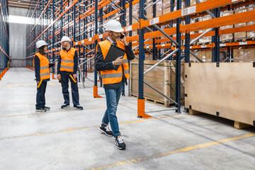 Warehouse manager using smartphone and holding clipboard is walking in a warehouse while workers are checking inventory