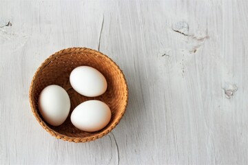 White eggs in a wicker basket on a white wooden background.
