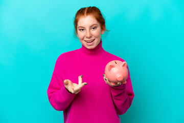 Young caucasian reddish woman holding a piggybank isolated on blue background with shocked facial...