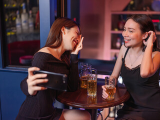 Two young Asian women laughing and taking a selfie while enjoying beer at a bar. A lively and candid moment of friendship, fun, and nightlife.