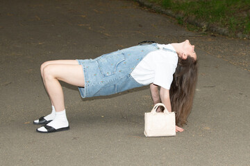 A person performs an unusual bridge pose on a park path, wearing a denim overall and carrying a...
