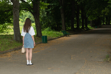 A young woman dressed in a casual outfit pauses on a quiet park path. Sunlight filters through the...