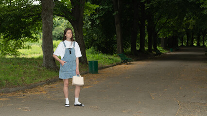 Fototapeta premium A young woman stands confidently in denim overalls, holding a small bag, surrounded by lush greenery in a peaceful park during the day. She enjoys the warm sun and fresh air