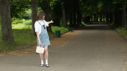 A young woman wearing a denim dress and white shirt stands on a quiet, sun-dappled path in a park,...
