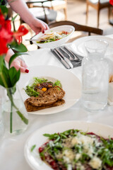 Person serving food at a restaurant table with plates, cutlery, water jug, and a floral centerpiece. Elegant indoor dining scene showcasing hospitality and gourmet presentation.