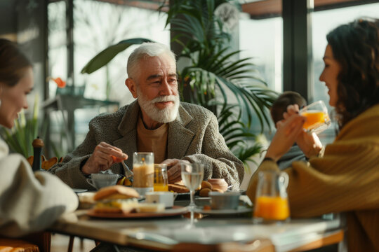 Senior man enjoying breakfast with family in a sunlit cafe, conversation, and laughter.