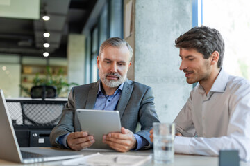 Mature businessman using a digital tablet to discuss information with a younger colleague in a...