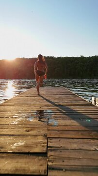 Slow motion shot of a girl jumping into the forest lake from a wooden ponton. Woman running on a wooden pier and jumping into the lake at sunset. Lens flare, top shot, aerial drone shot. Summer vibes.