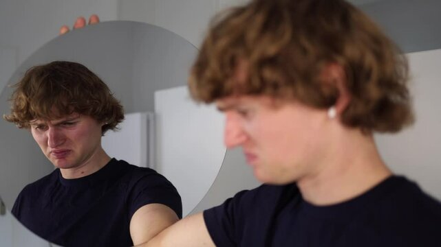 Young man looks at his reflection with a twisted face full of confusion and emotional pain as if asking why through clenched emotions in a raw moment of inner struggle