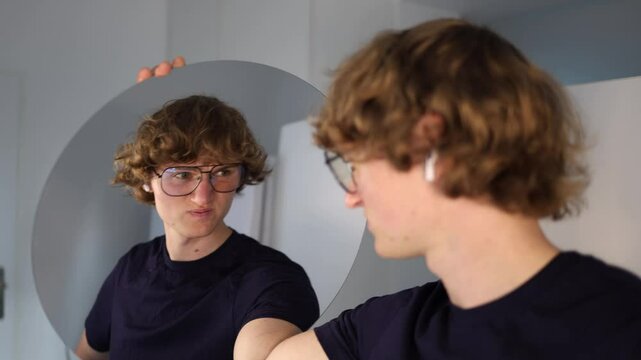 Young man clenches his jaw and lowers his head with a stiff posture while facing the mirror radiating an emotion of discomfort and awkward tension in a quiet self-conscious moment