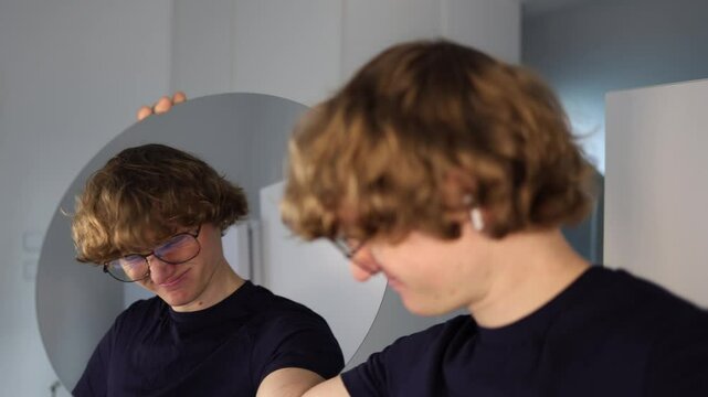Young man lowers his head and winces slightly while glancing at his reflection with a twisted smile showing a raw emotion of shame mixed with reluctant amusement