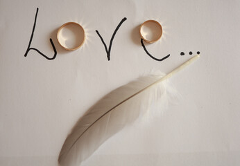 wedding rings forming the word love with a black marker on white paper, next to a feather