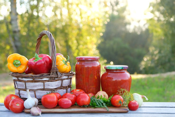 Glass jars with preserved homemade vegetables and ripe tomatoes, peppers, onions, garlic in basket on table outdoor, nature background. harvesting, assorted of raw vegetables for healthy vitamin food