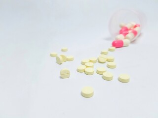 Close-up of yellow round tablets and pink-white capsules scattered on a white surface, symbolizing...