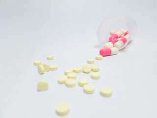 Close-up of yellow round tablets and pink-white capsules scattered on a white surface, symbolizing...