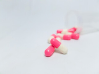 Blurry photo of pink and white capsules spilling from a transparent bottle on a white background,...
