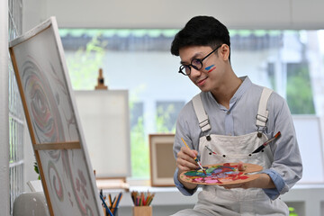 A Young Asian artist holding a colorful palette and paintbrush, thoughtfully blending paints in a...