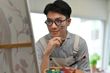 Young Asian male artist in overalls intently studies his canvas while holding a paintbrush and...