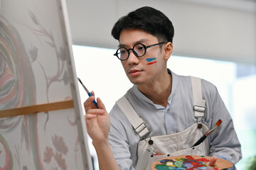 An Asian male artist with paint on his face confidently paints on canvas while holding a colorful...