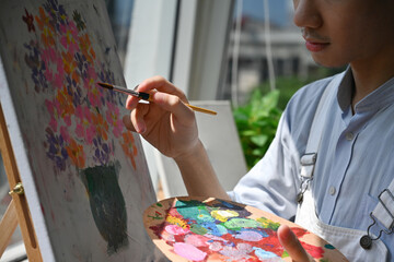 Close-up of an artist's hand painting colorful flowers on a canvas, holding a palette filled with...