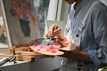 Close-up of a young artist hands mixing colors on a wooden palette, preparing paint for a floral...