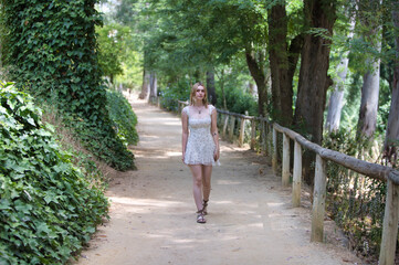 A young, beautiful blonde woman wearing a short floral print dress walks among trees along a path...