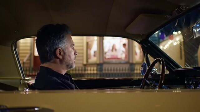 Mature man driving a vintage car at night looking focused through the windshield with city lights reflecting on it