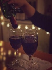 Hand pours red wine into glasses on a wooden table. Warm, dim lighting creates a cozy, intimate bar or restaurant ambiance.