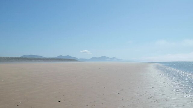 Scenic deserted sandy beach at Dinas Dinlle, Wales, UK with a light mist blowing over the sand and mountains under a bule sky on the horizon.